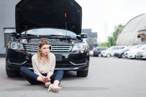 upset businesswoman sitting near broken auto with smartphone in hands, car insurance concept